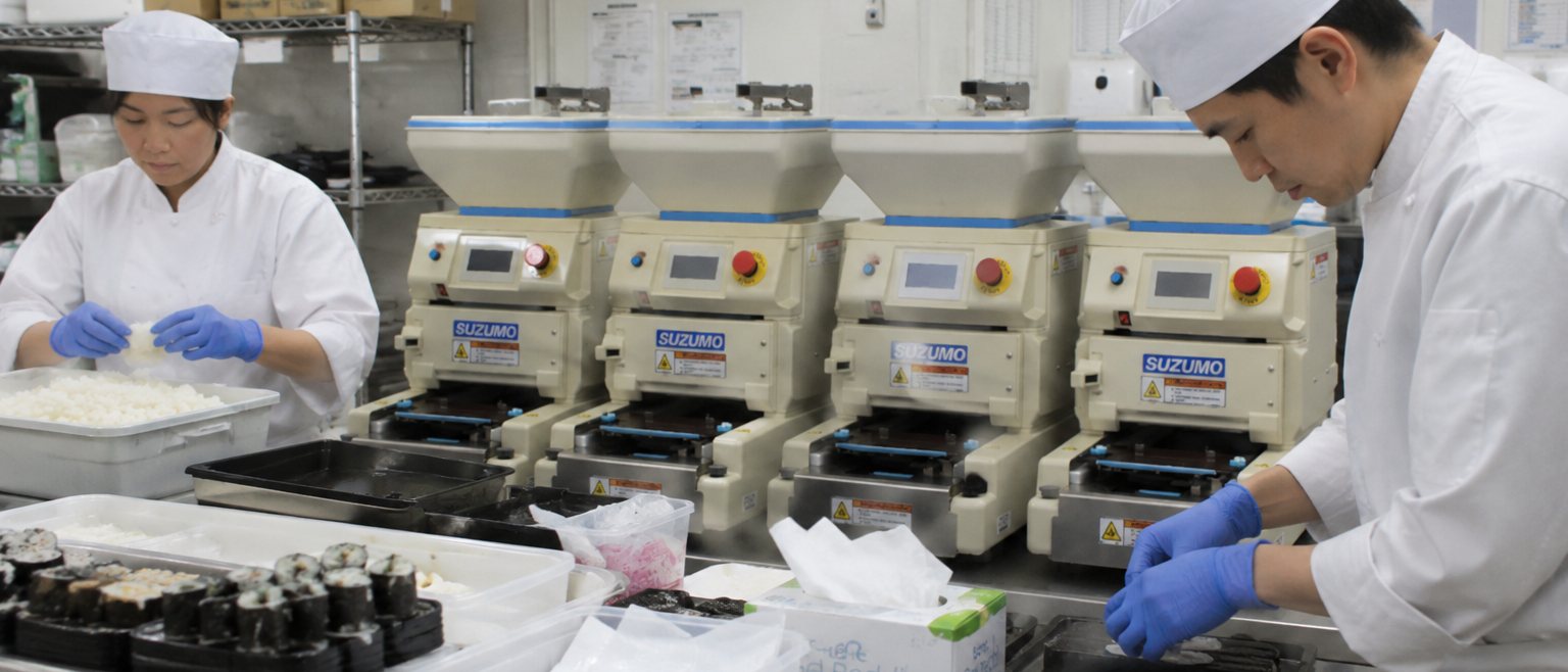Sushi chefs working with four Suzumo machines - trays of rolls in production